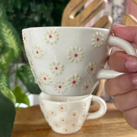 White Blossom Whisper mug with floral pattern held by a hand on a wooden surface with blurred greenery in the background