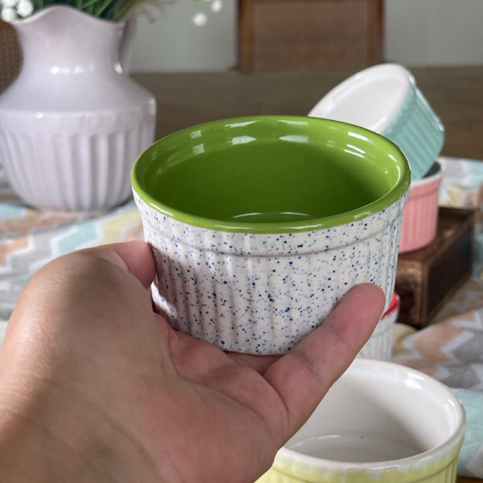 Hand holding a small ceramic pot with a green rim on a table with other pots and a vase in the background.