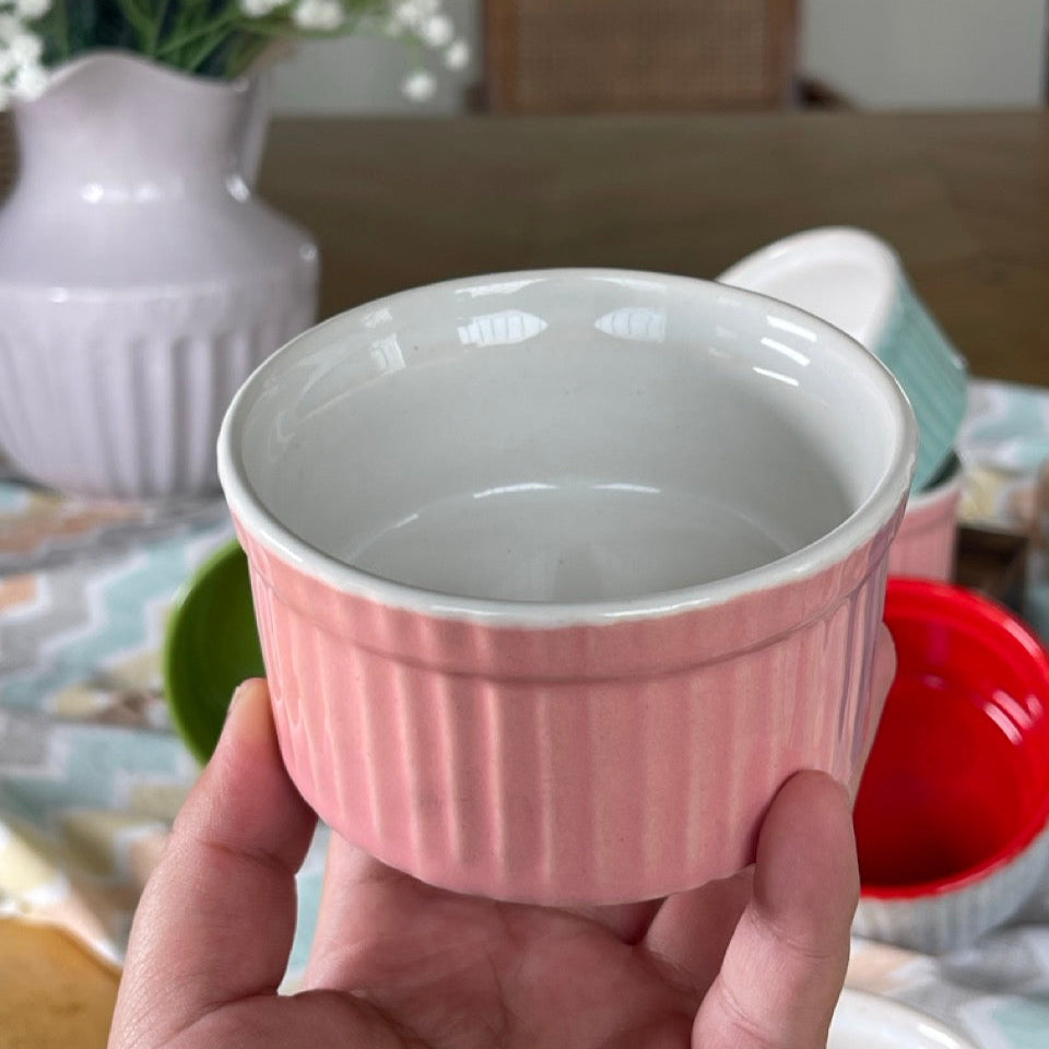 Hand holding a small pink ceramic ramekin with a blurred background of a table and flowers.