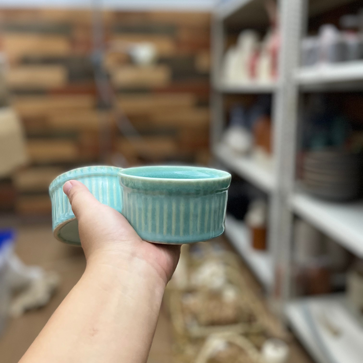Hand holding a small pastel teal blue ceramic bowl with a striped pattern in a blurred indoor setting.
