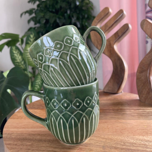 Green ceramic mugs with Kolam patterns stacked on a wooden surface with a plant in the background