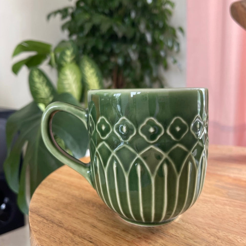 Green ceramic mug with decorative Kolam patterns on a wooden surface, with a blurred indoor background.