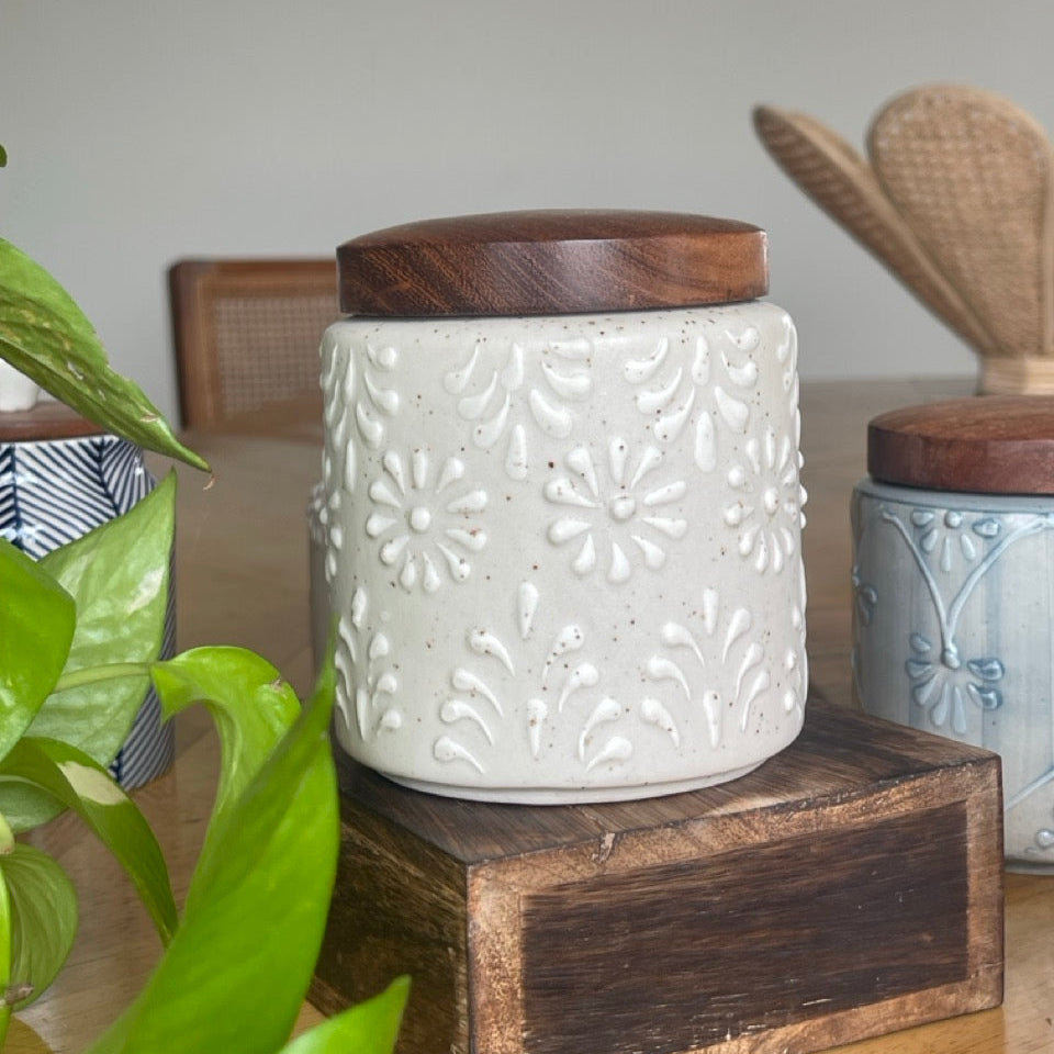 White ceramic jar with wooden lid on a wooden block, surrounded by greenery and other jars.