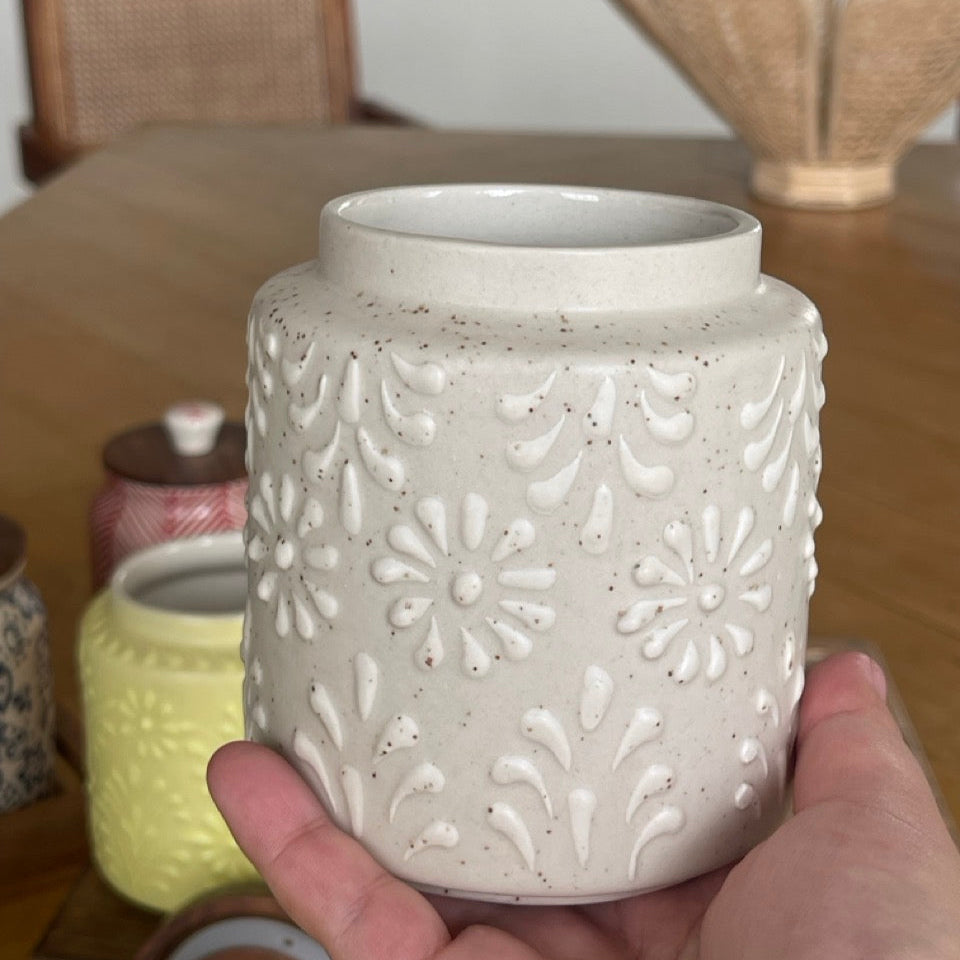 Hand holding a textured ceramic vase with floral patterns on a blurred indoor background