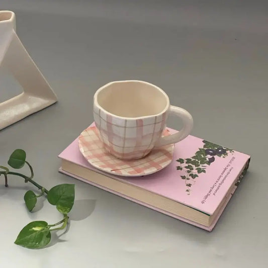Pink checkered ceramic mugs and saucer on a pink book with a plant on a gray surface