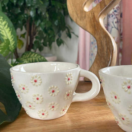 Two ceramic cups with floral patterns on a table with a plant in the background.