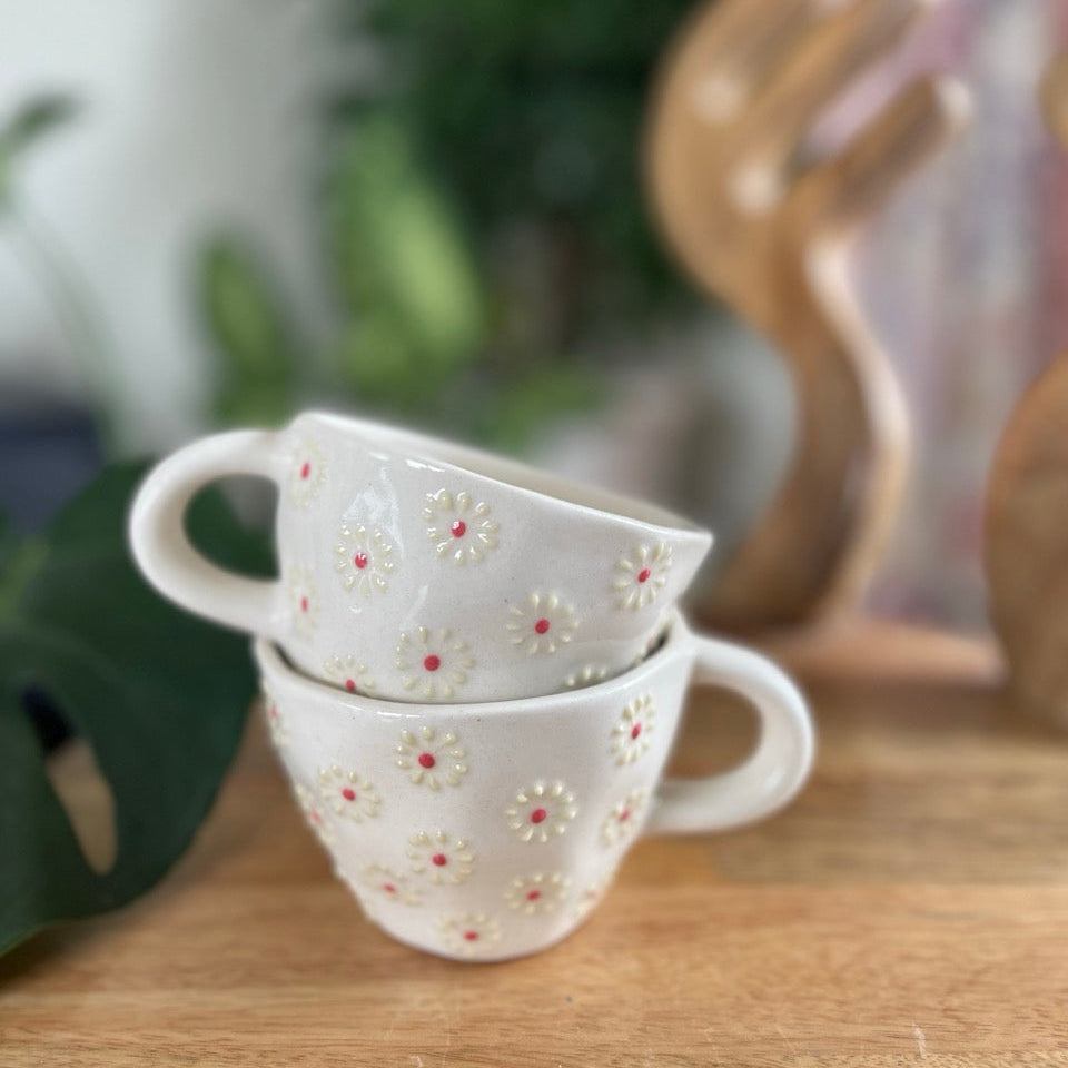 Two white ceramic mugs with floral patterns stacked on a wooden surface, blurred greenery in the background