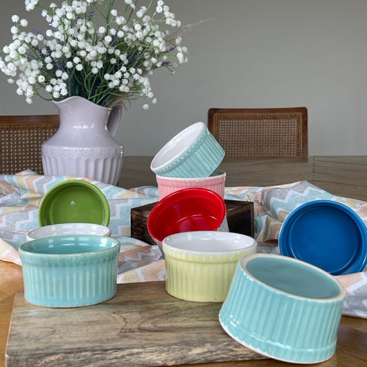 Colorful ceramic ramekins on a wooden surface with a vase of white flowers in the background.