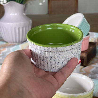 Hand holding a small ceramic pot with a green rim on a table with other pots and a vase in the background.
