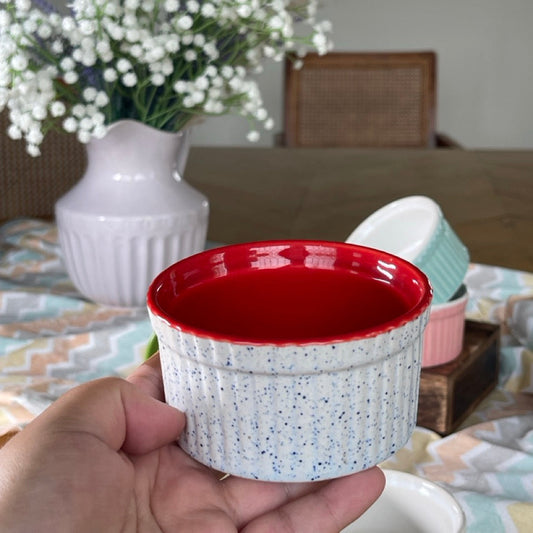 Speckled ceramic ramekin with red interior held in front of a blurred dining table setting.