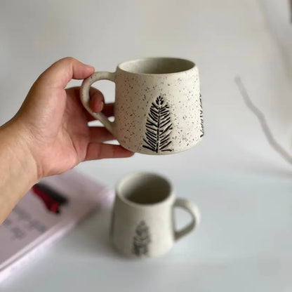 Hand holding a ceramic Evergreen glyph mug with a leaf design on a light background