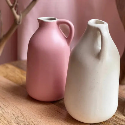 Two ceramic Jugscapes vases, one pink and one beige, on a wooden surface with a blurred pink background.