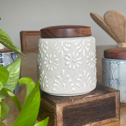 White ceramic jar with wooden lid on a wooden block, surrounded by greenery and other jars.