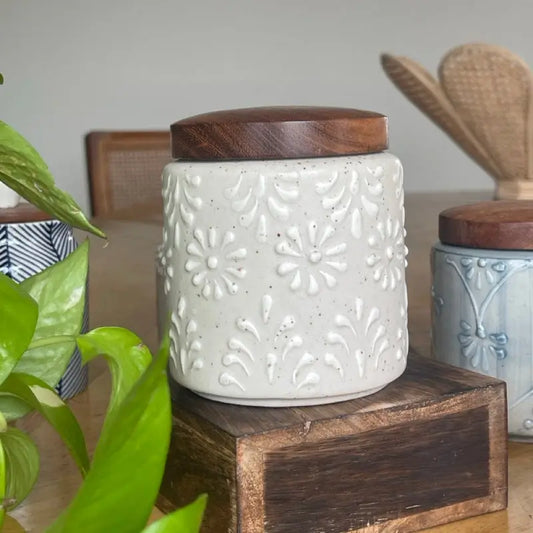 White ceramic Keepsake jar with wooden lid on a wooden block, surrounded by greenery and kitchen items.