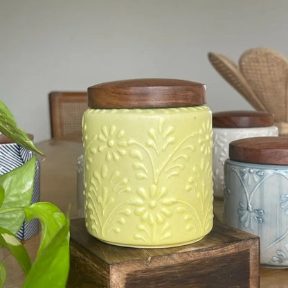 Yellow Keepsake ceramic jar with wooden lid on a wooden surface, surrounded by other jars and a plant.