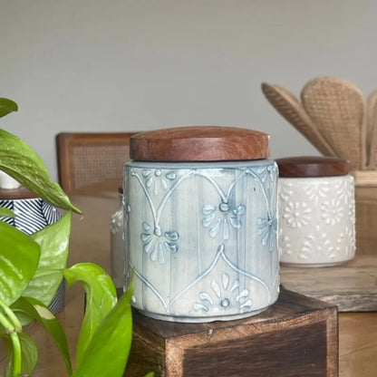 Keepsake ceramic jar with wooden lid on a wooden surface, surrounded by greenery.