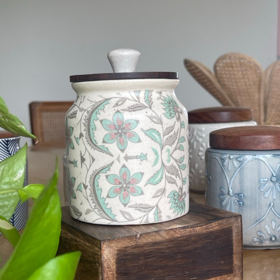 Decorative ceramic jar with floral design on a wooden stand, surrounded by other jars and plants.