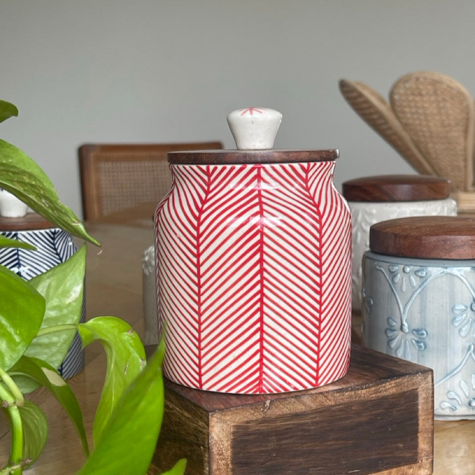 Ceramic jar with red and white pattern on a wooden stand, surrounded by plants and other items.