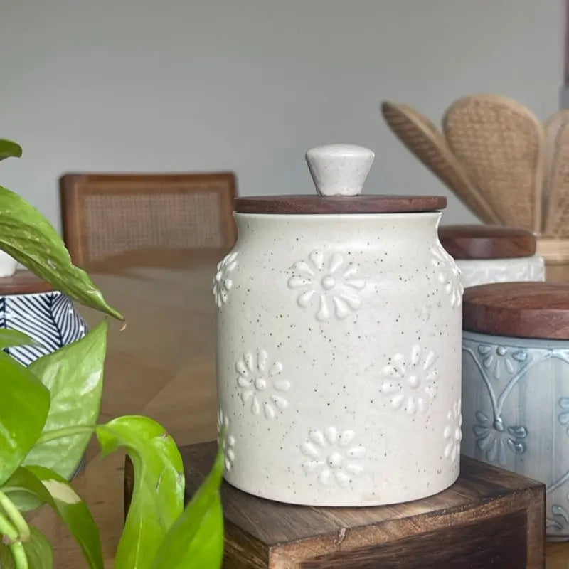 White Keepsake ceramic jar with wooden lid on a wooden surface, with a blurred indoor setting in the background.