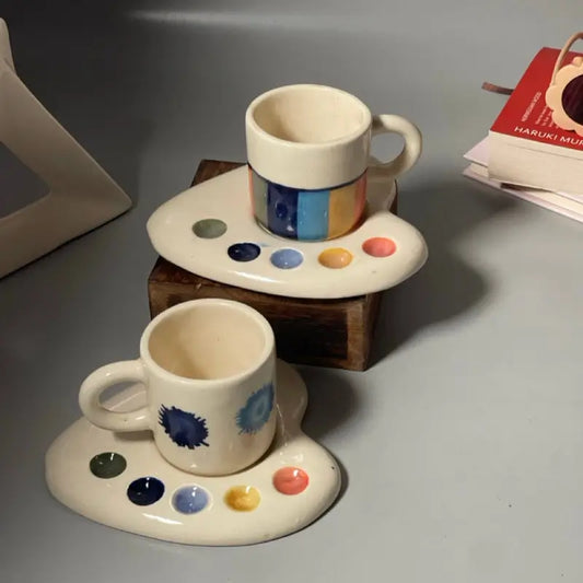 Two Palette shaped ceramic cups with artistic designs on saucers on a gray surface.