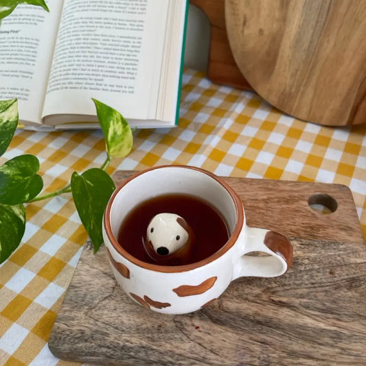 Pawprint Mug with hidden dog figurine Tea cup on a checkered tablecloth with an open book and wooden cutting board.