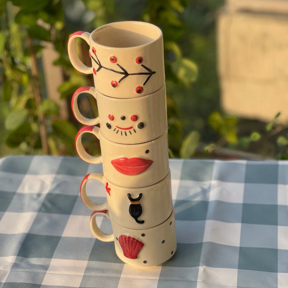 Stack of decorative ceramic mugs with facial expressions on a checkered tablecloth.
