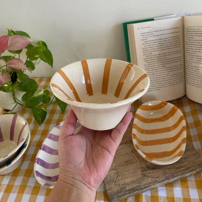 Hand holding a ceramic bowl with orange stripes on a checkered tablecloth with books and plants.