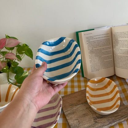 Hand holding a striped ceramic bowl on a table with a book and plant in the background
