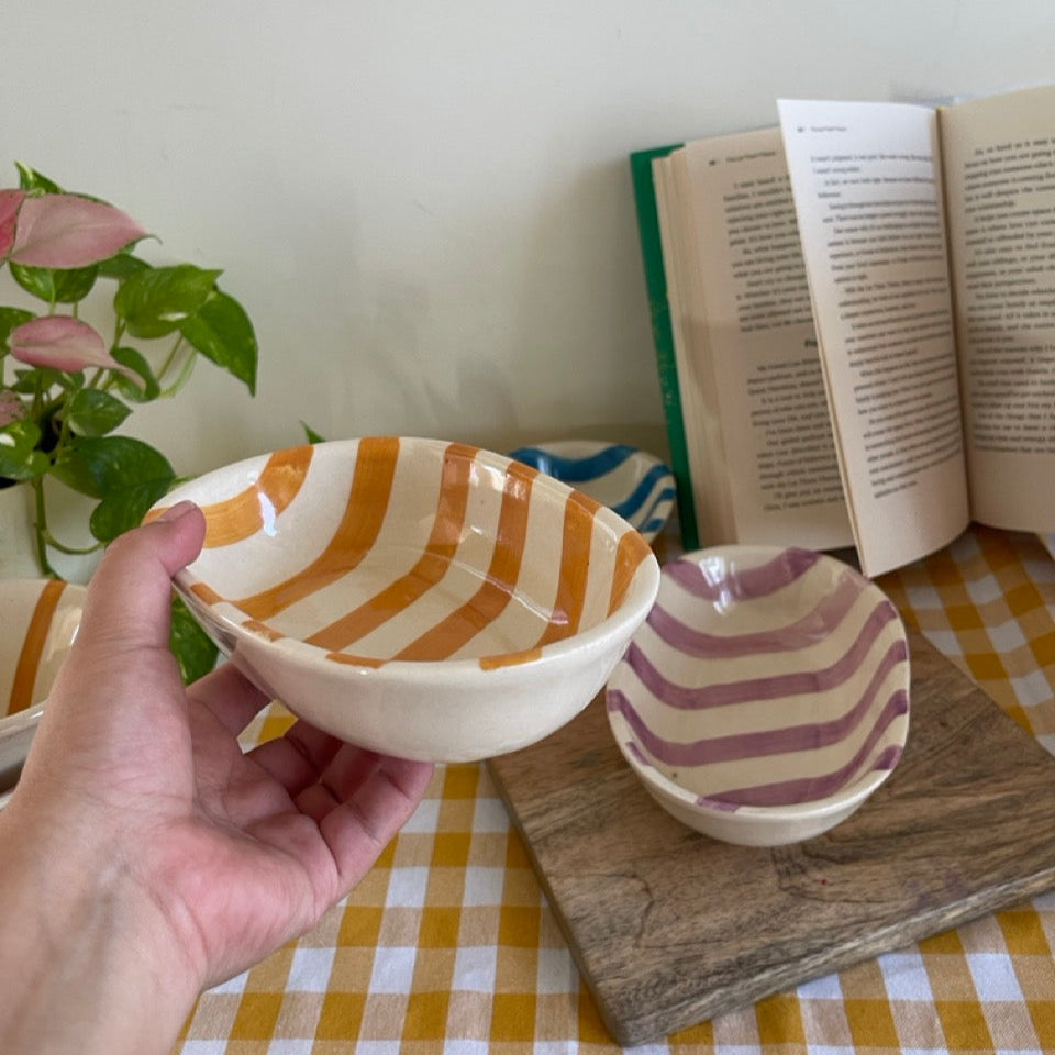 Hand holding a ceramic bowl with striped pattern on a checkered tablecloth with another bowl and book in the background.