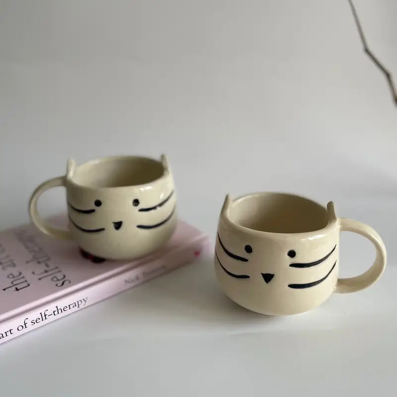 Two cat-shaped ceramic mugs on a surface with a book in the background.